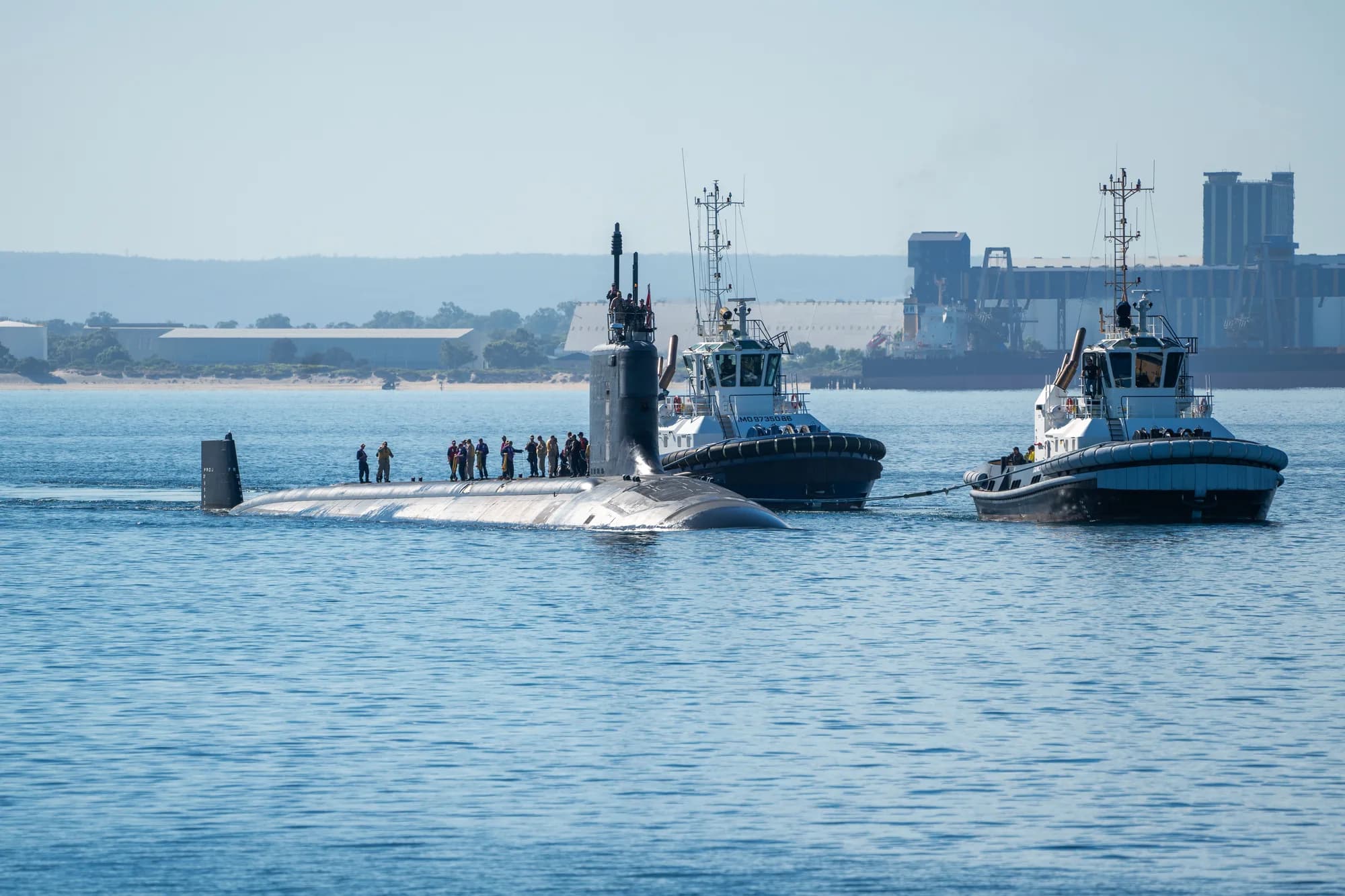 A United States Virginia-class submarine under tow in Cockburn Sound with industrial port infrastructure behind